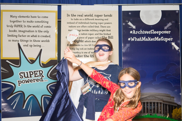 Zwei junge Mädchen in Superheldenkostümen posieren vor einem blauen Vorhang auf einem Schulereignis, eine hält ein Tuch und beide tragen Masken.