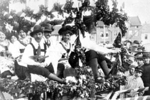 Eine Gruppe von Menschen in traditioneller bayerischer Tracht fahrt auf einem mit Bl├Ąttern geschm├╝ckten Schwimmwagen in einem Umzug, einige halten Musikinstrumente, mit Geb├Ąuden und einem klaren Himmel im Hintergrund, dargestellt in Schwarz-Wei├č.