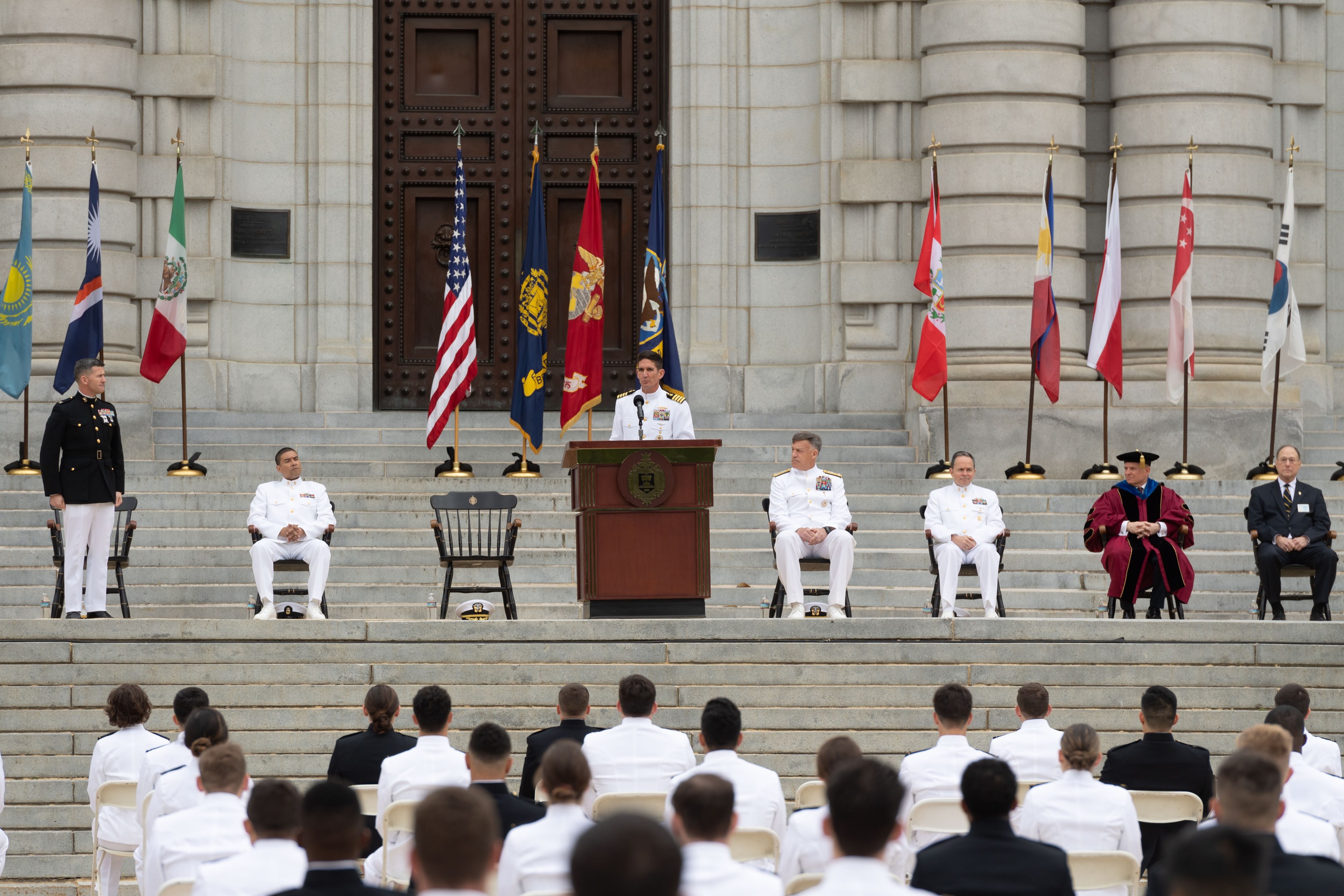 Gruppe von Menschen in weißen Uniformen auf Treppe eines Gebäudes mit Podium, flankiert von Fahnen, vor einem großen Gebäude mit einer Tür während einer offiziellen Zeremonie.