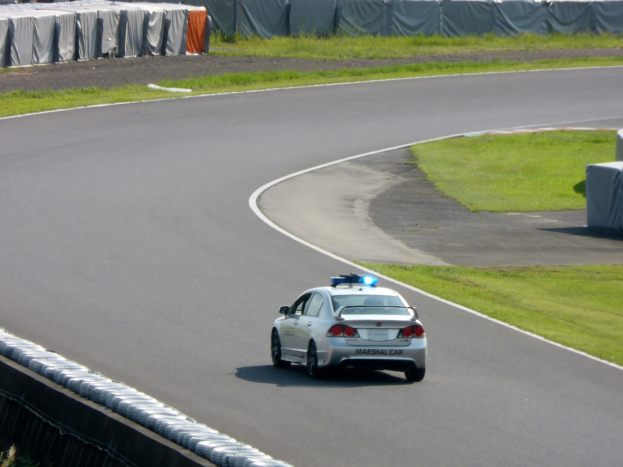 Polizeiauto auf einer Rennstrecke mit Gras an beiden Seiten und Zuschauern im Hintergrund.