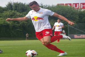 Ein Profifußballer kickt einen Ball auf einem Rasenfeld mit Bäumen und einem klaren blauen Himmel im Hintergrund, in der Nähe eines Schilds mit der Aufschrift "RB Leipzig."