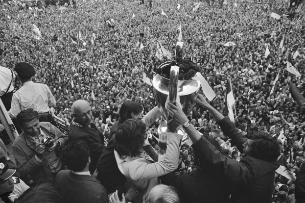 Schwarzes und weißes Bild einer Gruppe von Menschen, die einen Pokal hochhalten, während einige Kameras und Fahnen halten, vor einer großen Menge.
