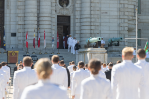 Eine Gruppe von Menschen in weißen Uniformen und Mützen steht vor einem Gebäude mit Säulen und einer Tür, mit Flaggen, einem Podium mit Mikrofon, Stufen und Kanonen im Hintergrund, was auf eine Abschlussfeier der Marineakademie hindeutet.