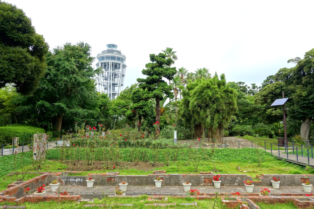 Ein Park mit einem Turm im Hintergrund, umgeben von grünem Gras, Pflanzen und Bäumen, mit verstreuten Blumentöpfen, einer Straße mit Geländern und einem Pfahl mit einer Tafel, unter einem sichtbaren Himmel.