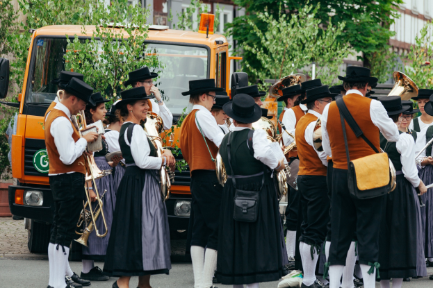 Gruppe von Menschen in traditioneller bayrischer Tracht, die auf der Straße spielen und Taschen tragen, mit Bäumen, Gebäuden und einem geparkten Fahrzeug im Hintergrund.