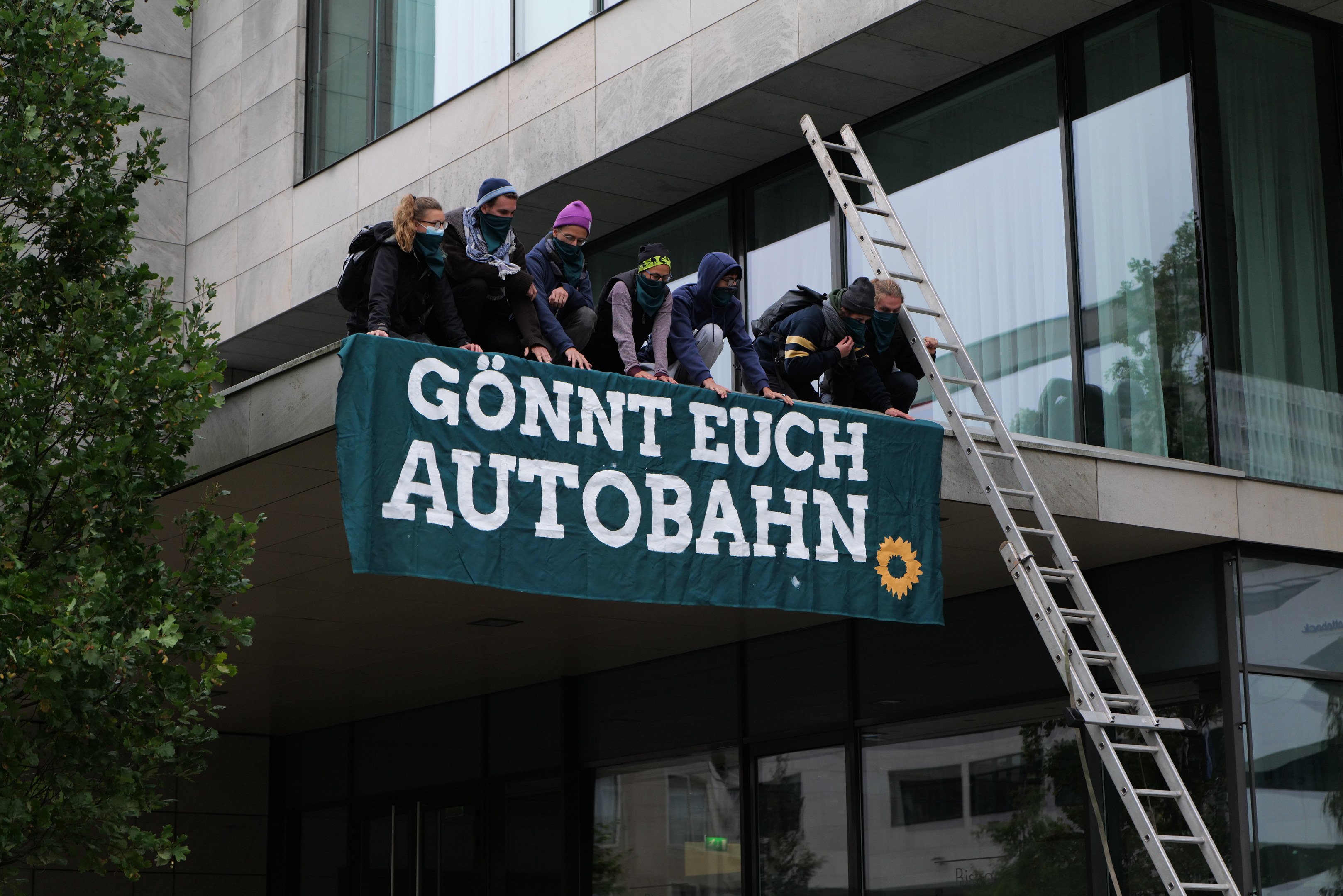 Menschen auf einem Gebäude mit einem Transparent, links ein Baum und Glasfenster im Hintergrund, scheinbar protestierend für die deutsche Regierung.