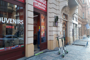 Eine Stadtstraße mit einer Reihe geparkter Scooter, gesäumt von Gebäuden mit Fenstern, Türen und Texttafeln, einem Geländer und Bäumen vor einem klaren blauen Himmel im Hintergrund.