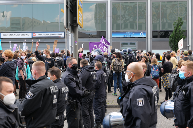 Eine große Gruppe von Menschen vor einem Gebäude, einige mit Schildern und Helmen, mit einem Pfahl mit einer Schildertafel im Vordergrund und einem Baum im Hintergrund, scheinbar protestierend.
