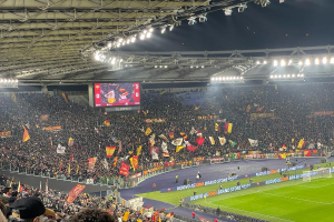 Eine große Menschenmenge in einem Stadion mit schwenkenden Fahnen, Gras, einem Netz und Bannern mit Text auf dem Boden, viele Zuschauer in den Rängen, einige mit Mützen, ein Bildschirm im Hintergrund und beleuchtete Deckenlampen, die eine Atmosphäre der Aufregung und Vorfreude auf ein Fußballspiel schaffen.