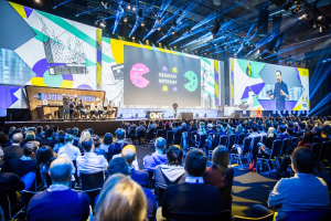 Eine große Menschenmenge sitzt auf Stühlen vor einer Bühne bei der Deutschen Internet-Konferenz 2019, mit einem Tisch voller Gegenstände links und Bildschirmen und Lichtern im Hintergrund.