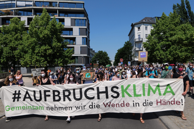 Gruppe von maskierten Menschen mit einem Banner 'Aufbruchsklima' vor einem Gebäude mit Bäumen und wolkenlosem Himmel.
