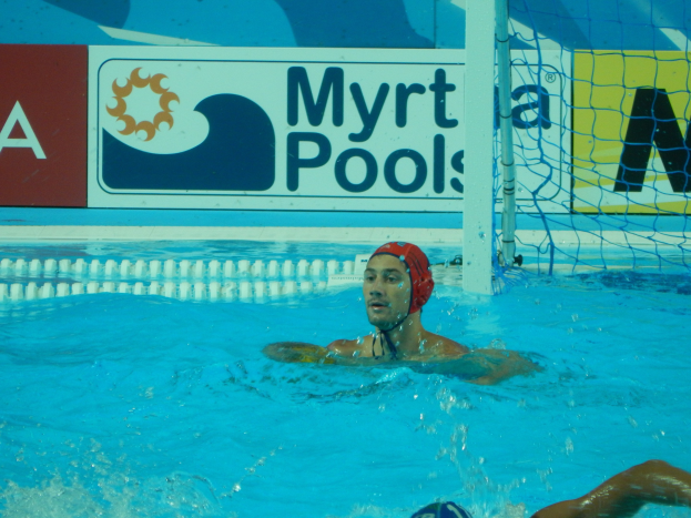 Wasserballspieler in Bewegung in einem Pool, trägt eine Mütze, mit einem Netz auf der rechten Seite und einer Werbetafel im Hintergrund.