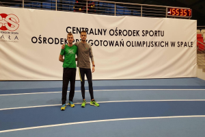 Zwei Männer stehen auf einem Tennisplatz mit einem Banner im Hintergrund, das 'Olympische Spiele 2019 - Ostrodek vs Spale' lautet, sowie einer Digitalanzeigetafel, einem Geländer und ein paar Stühlen.