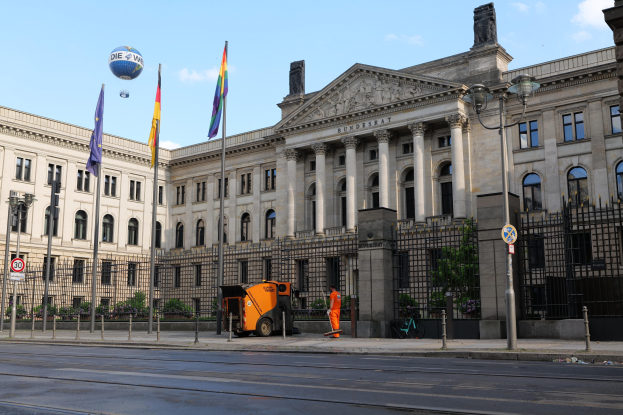Großes modernes Gebäude mit zahlreichen Fenstern, als Bundestag in Berlin, Deutschland identifiziert, umgeben von Straßeninfrastruktur und Fußgängern, mit Flaggen, Bäumen und einem bewölkten Himmel im Hintergrund.