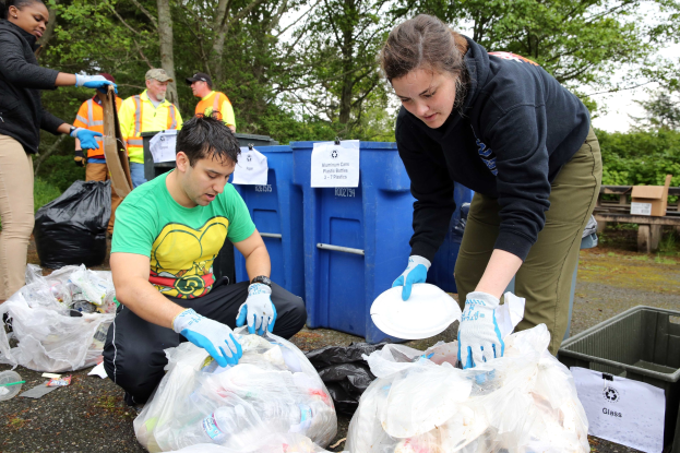 Eine Gruppe von Menschen, die Müll in einem Park sammeln, mit einem Mann und einer Frau in der Mitte, die Handschuhe tragen und Schilder halten, umgeben von Plastikabdeckungen, Flaschen und anderen Gegenständen auf dem Boden, einem Müllcontainer und einer Holzbank rechts und Bäumen mit einem klaren blauen Himmel im Hintergrund.