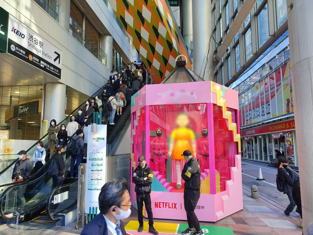 Eine Gruppe von Menschen steht vor einem Gebäude mit Glaswänden, Säulen und Texttafeln, einige tragen Masken, während eine Rolltreppe mit Menschen zu sehen ist. Eine rosafarbene Box in der Mitte zeigt Schaufensterpuppen und trägt die Aufschrift "Netflix's new Netflix booth in Tokyo."