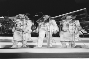 Drei Frauen singen in Microphone auf der Bühne mit Musikern im Hintergrund, Schwarz-Weiß.