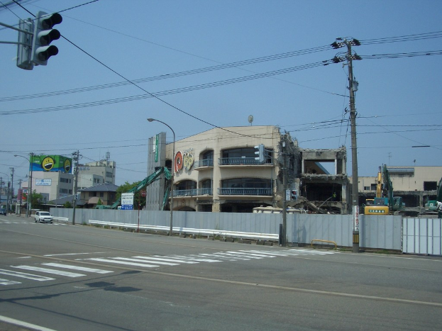 Eine Stadtstraße mit Fahrzeugen, Strommasten, Laternen, einer Ampel auf der linken Seite, flankiert von Gebäuden, Bäumen und dem Himmel, mit einem teilweise abgerissenen Gebäude im Vordergrund.