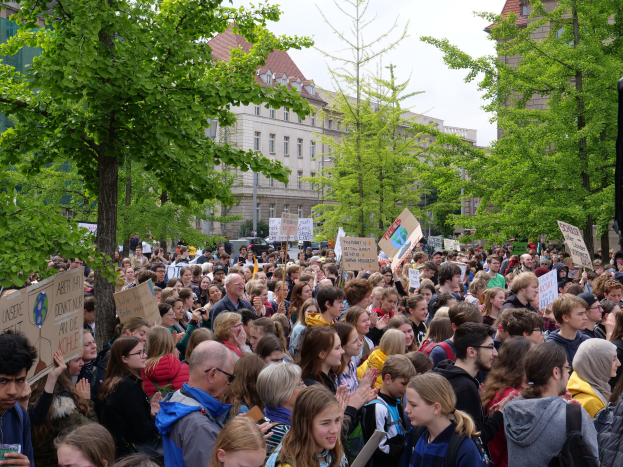 Eine große Menschenmenge protestiert vor einem Gebäude in Berlin, hält Schilder, mit Bäumen, Fahrzeugen, einem Lautsprecher und Himmel.