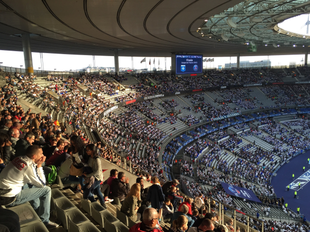 Eine große Menschenmenge in einem Stadion schaut ein Fußballspiel zu, mit einer Bühne rechts, Fahnen, Stangen, einem Bildschirm und dem Allianz Stadion in München im Hintergrund.