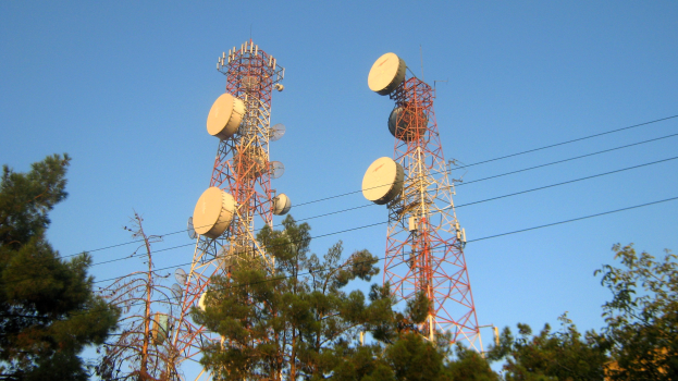 Zwei Mobilfunkmasten mit Antennen, umgeben von Bäumen und Drähten, unter einem klaren blauen Himmel.
