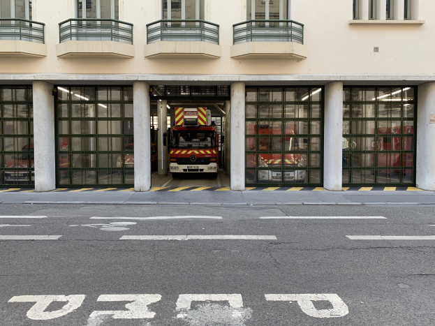 Feuerwache mit einem Feuerwehrauto davor, das Fenster, Geländer, Säulen, Glastüren und eine Straße am Boden zeigt.