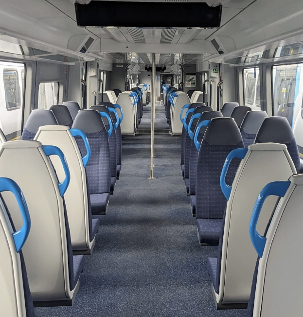 Innenseite eines Zuges mit blauen und weißen Sitzen, hohen Pfosten, Fenstern und hellen Deckenleuchten.