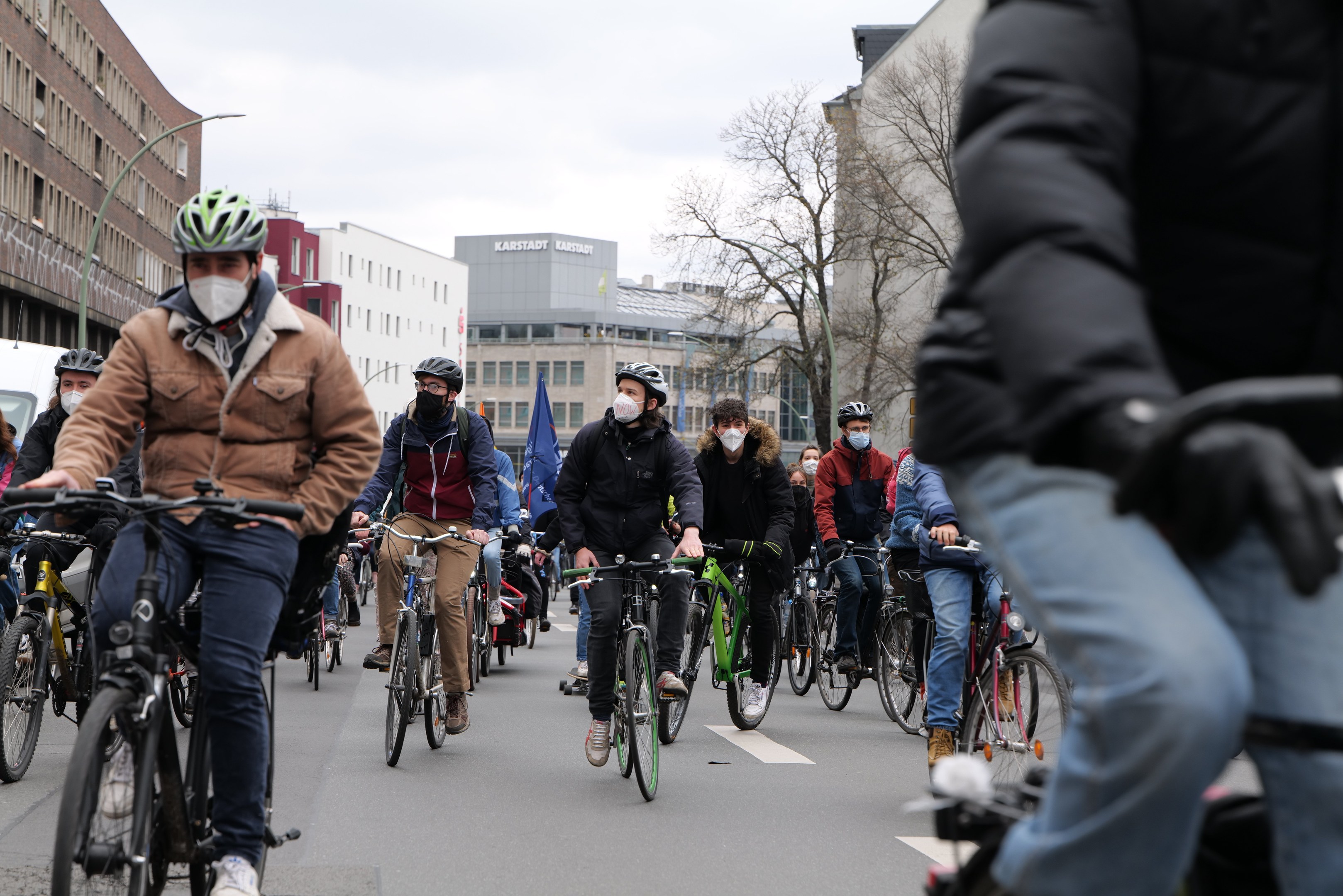 Eine Gruppe von Menschen in Helmen und Handschuhen fährt Fahrräder auf einer von Bäumen gesäumten Straße in Berlin, mit Gebäuden und einem geparkten Fahrzeug im Hintergrund.