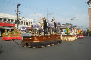 Ein Paradewagen mit Menschen darauf, umgeben von Gebäuden, Strommästen mit Drähten, Laternenmästen, Bäumen und einem Uhrenturm im Hintergrund bei bewölktem Himmel.