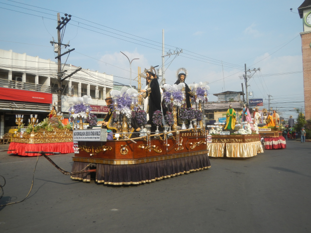 Ein Paradewagen mit Menschen darauf, umgeben von Gebäuden, Strommästen mit Drähten, Laternenmästen, Bäumen und einem Uhrenturm im Hintergrund bei bewölktem Himmel.