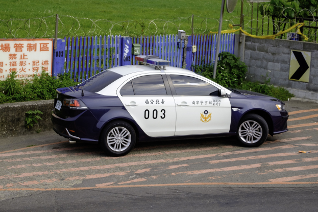 Polizeifahrzeug am Straßenrand mit umgebender Vegetation, einem Zaun, einem Schild, einer Wand, einem Strommast, einer Satellitenschüssel und Bäumen.