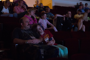 Eine Gruppe von Menschen sitzt in einem Kinosaal mit einem Bildschirm auf der rechten Seite und einer Wand im Hintergrund.