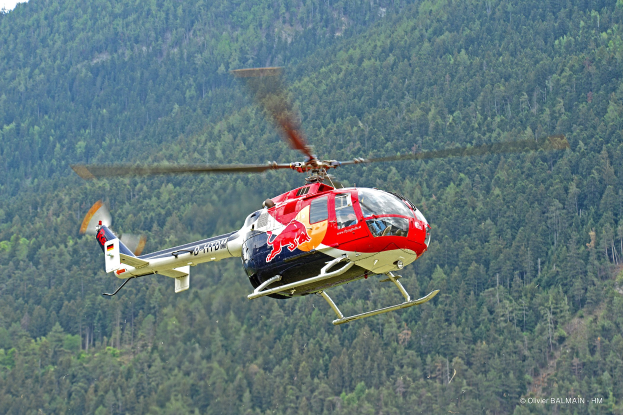Roter Bullen-Hubschrauber fliegt über einen grünen Wald mit Bäumen auf Hügeln und einem klaren blauen Himmel.