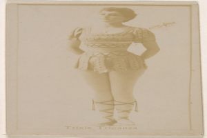 Ein altes Foto einer Frau in einem hellfarbenen gemusterten Badeanzug, die Haare zu einem Dutt gebunden, steht und lächelt direkt in die Kamera mit ihren Händen vor der Brust verschränkt.