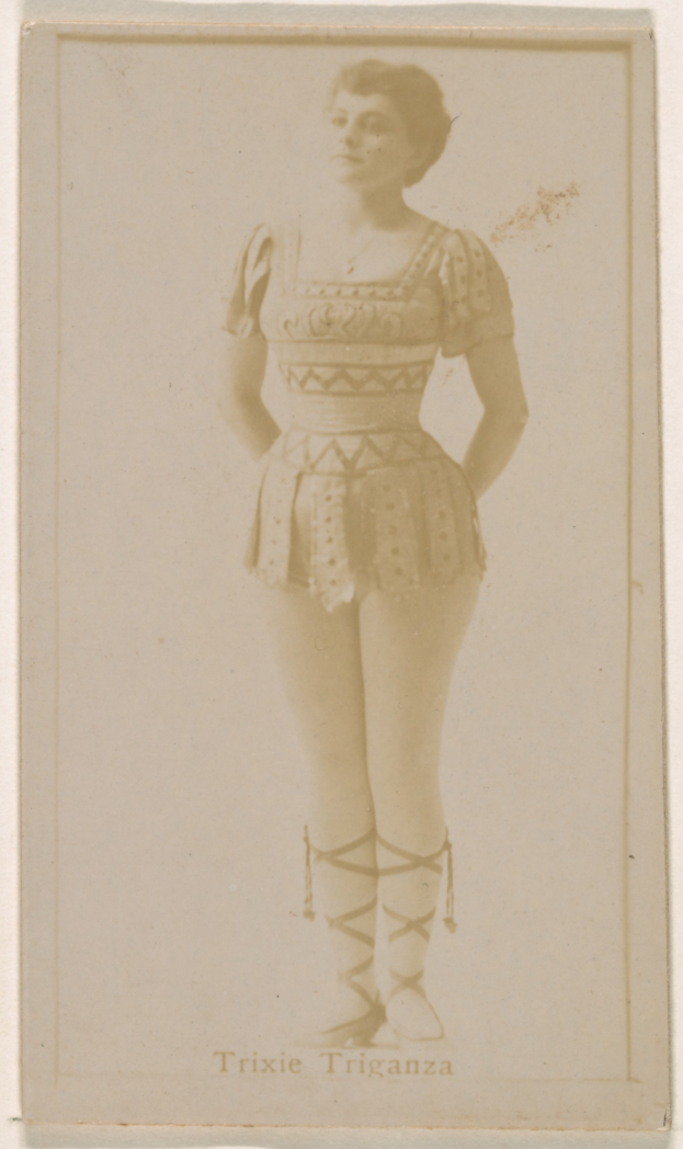 Ein altes Foto einer Frau in einem hellfarbenen gemusterten Badeanzug, die Haare zu einem Dutt gebunden, steht und lächelt direkt in die Kamera mit ihren Händen vor der Brust verschränkt.