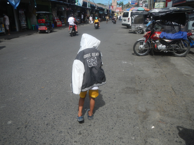 Ein junger Junge geht eine Straße entlang, die von Motorrädern und anderen Fahrzeugen sowie Gebäuden mit Schildern gesäumt ist, unter einem klaren blauen Himmel mit Bäumen und Masten im Hintergrund.