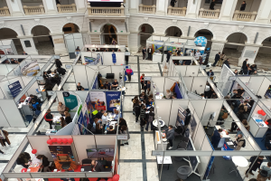 Große Besuchergruppe auf einer internationalen Fachmesse mit Ständen, Bannern, Tischen, Stühlen und Ballons in einem Raum mit Bogengängen, Säulen und einem Bildschirm im Hintergrund.