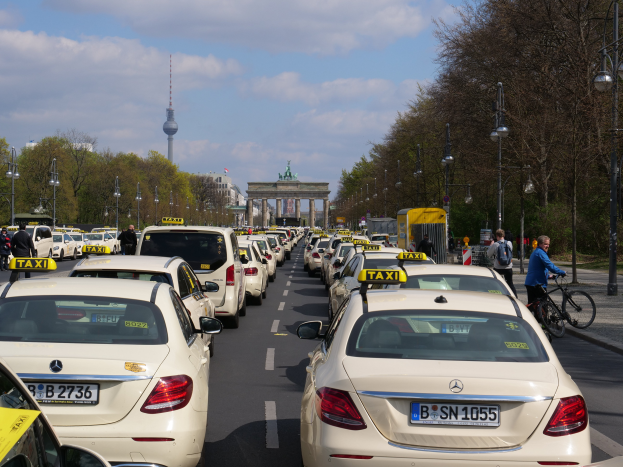 Eine lange Reihe von Taxis, die auf der Seite einer belebten Straße in Berlin, Deutschland, geparkt sind, mit Fahrzeugen, Radfahrern und Fußgängern, flankiert von Laternenpfählen, Bäumen und Gebäuden, einschließlich eines Bogens und eines Turms.