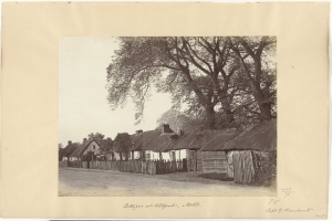 Schwarzes und weißes Foto eines Dorfes mit Reetdächern, einer Straße, einem Zaun, Bäumen und einem Himmel im Hintergrund, mit Text unten.