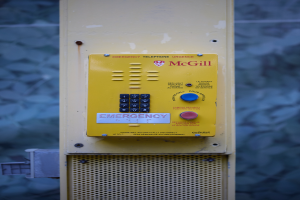 Ein gelbes Notruf-Telefon an einer Gebäudeseite angebracht, mit Tasten und Text zum Absetzen von Notrufen, vor einer Wand.