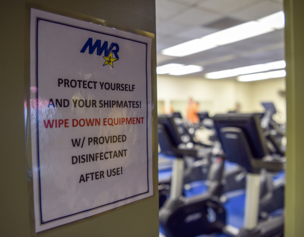 Ein Schild an einer Turnhallentür mit der Aufschrift "Schützen Sie sich und Ihre Schiffskameraden" mit Laufbändern im Hintergrund und Deckenleuchten, das eine leicht unscharfe, bewegungsähnliche Wirkung hat.