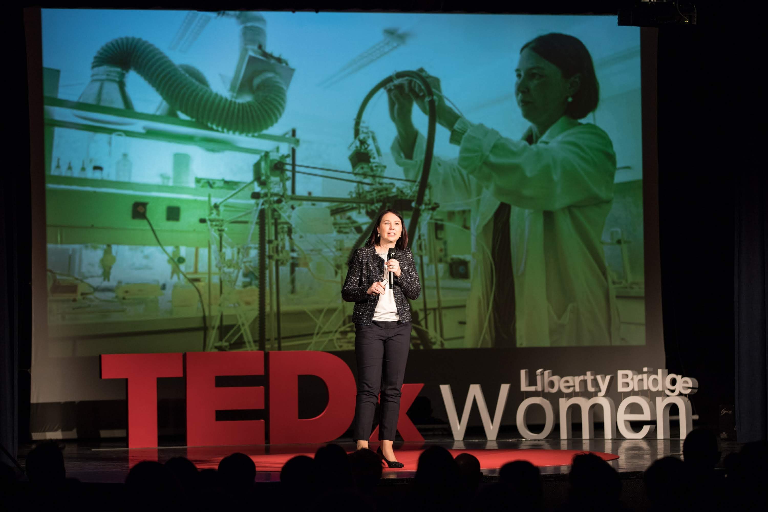 Frau auf einer Bühne mit Mikrofon, hinter ihr ein Display mit einem Bild einer Frau und Text zum TEDxWomen-Event.