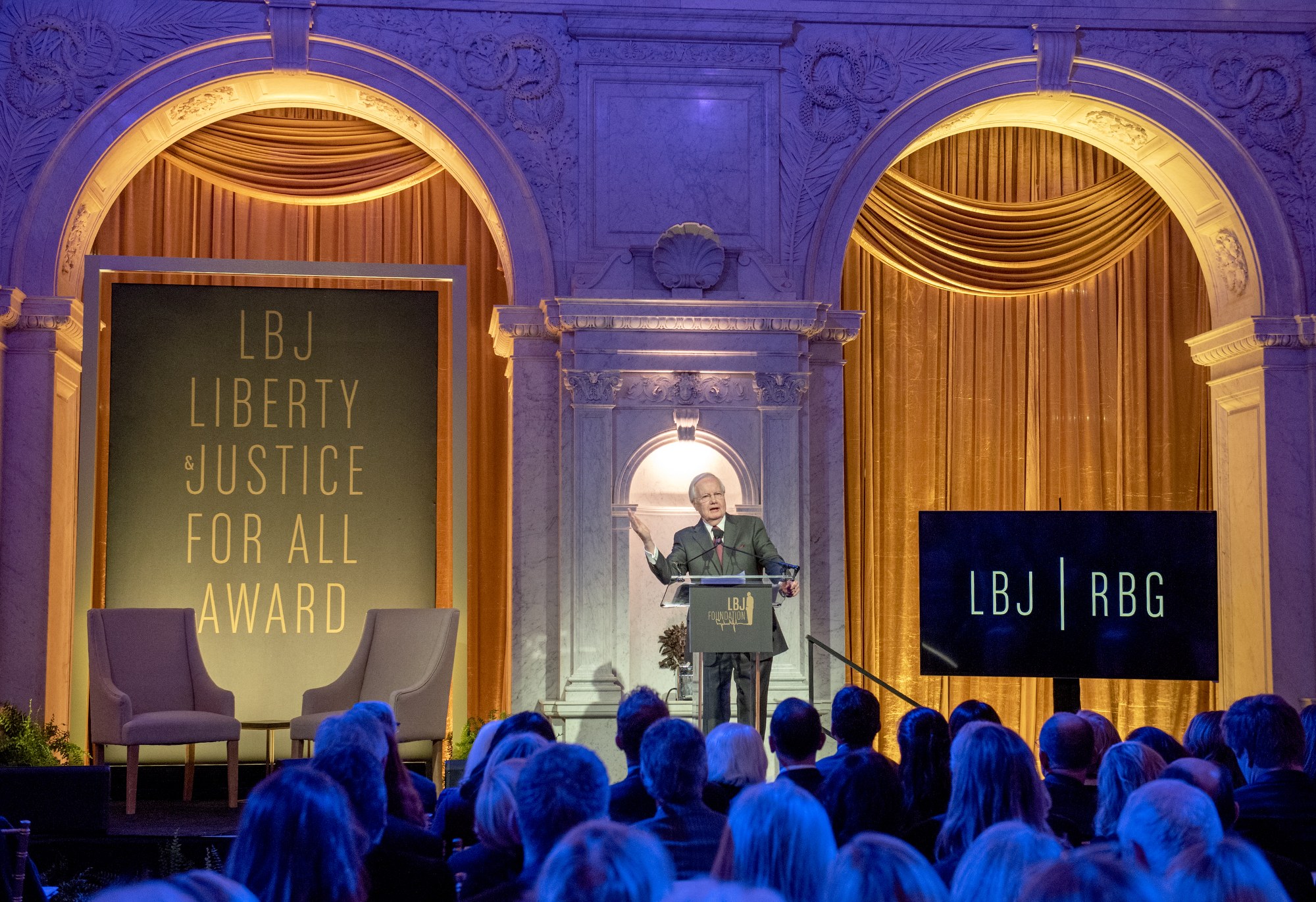 Ein Mann steht an einem Rednerpult mit einem Mikrofon, spricht zu einer sitzenden Menge, mit einer Tafel mit Text dahinter und Pflanzen und Vorhängen im Hintergrund.