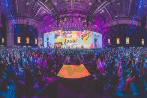 Eine große Menschenmenge auf Stühlen sitzend, die einer hell erleuchteten Bühne gegenüberstehen, umgeben von Bildschirmen, wahrscheinlich bei einem Online-Marketing-Rockstars-Event.