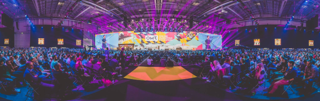 Eine große Menschenmenge auf Stühlen sitzend, die einer hell erleuchteten Bühne gegenüberstehen, umgeben von Bildschirmen, wahrscheinlich bei einem Online-Marketing-Rockstars-Event.