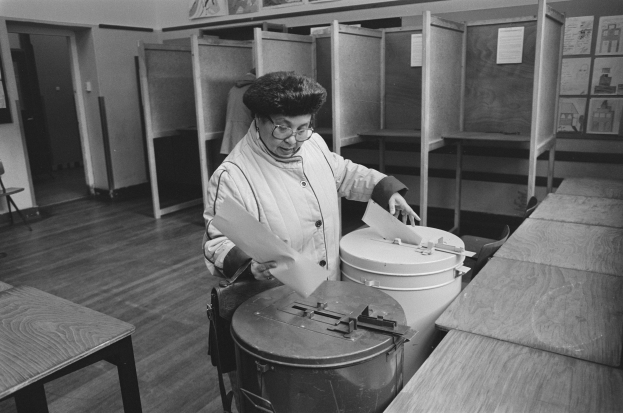 Eine Frau mit Brille hält Papiere in der Hand, während sie in einer Wahllokal wählt, mit Tischen, Bänken, Schränken, einer Tür, einem Stuhl und Plakaten im Hintergrund, alles in Schwarz-Weiß.