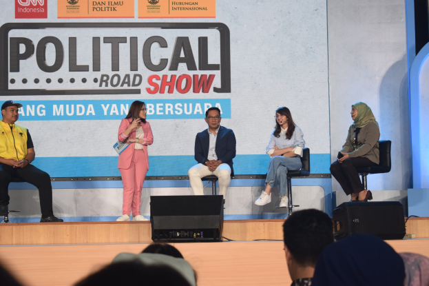 Eine Gruppe von Menschen sitzt auf einer Bühne mit einer Frau in der Mitte, die ein Mikrofon hält, einer Zuschauermenge im Vordergrund und einem Banner mit der Aufschrift 'Politischer Straßenumzug 2019' im Hintergrund.