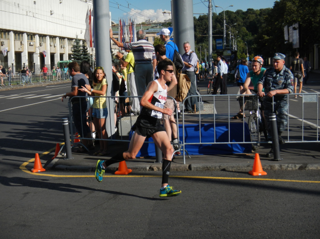 Ein Mann, der auf einer Stadtstraße bei einem Marathon läuft, umgeben von Zuschauern mit Fahrrädern, mit städtischer Infrastruktur und einem bewölkten Himmel.