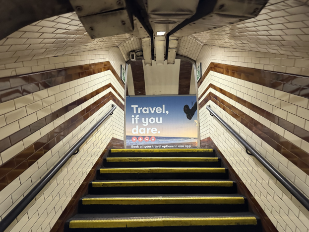 Eine Treppe in einer U-Bahn-Station mit einem "Reise, wenn du wagst"-Schild darüber, flankiert von Geländern und U-Bahn-Stationswänden im Hintergrund.