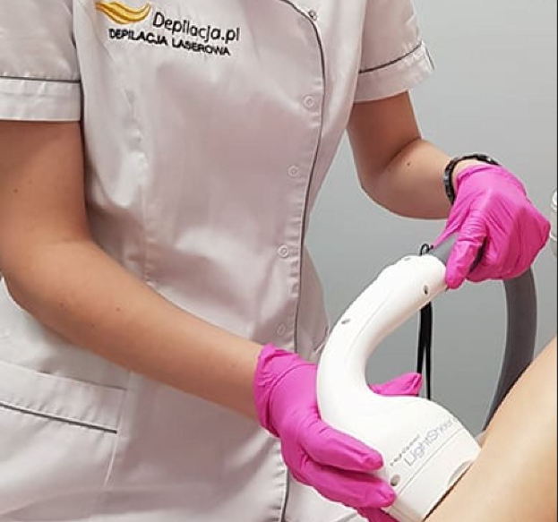 Eine Frau in einem weißen Hemd und pinken Handschuhen bedient eine weiße Laser-Haarentfernungsmaschine auf ihrer Haut, vor einer Wand stehend.